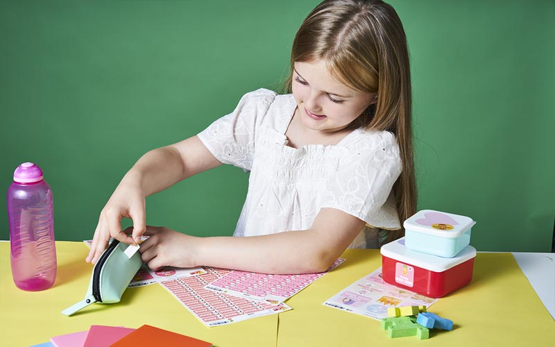 Une fille montrant à quel point les autocollants de nom My Nametags sont faciles à utiliser en les mettant sur une trousse à crayons